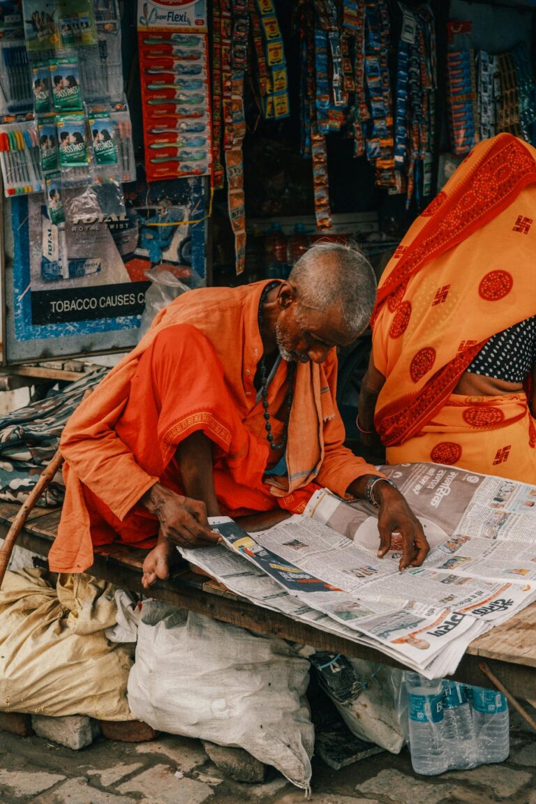 An elderly man in traditional attire reading a newspaper at a vibrant Indian market stall.