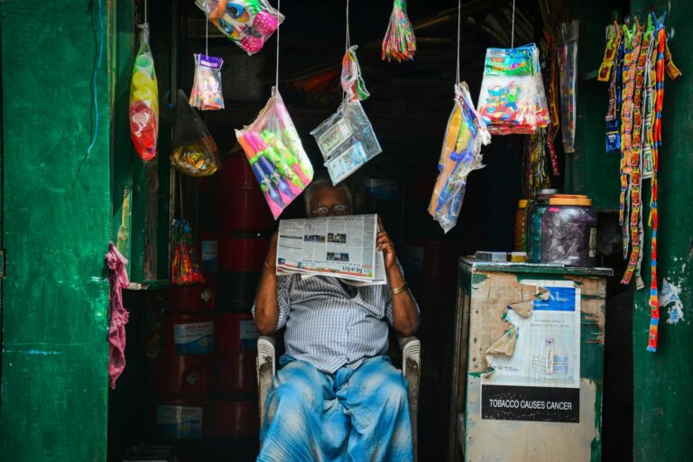 An elderly man reading a newspaper in a vibrant market shop filled with hanging goods.