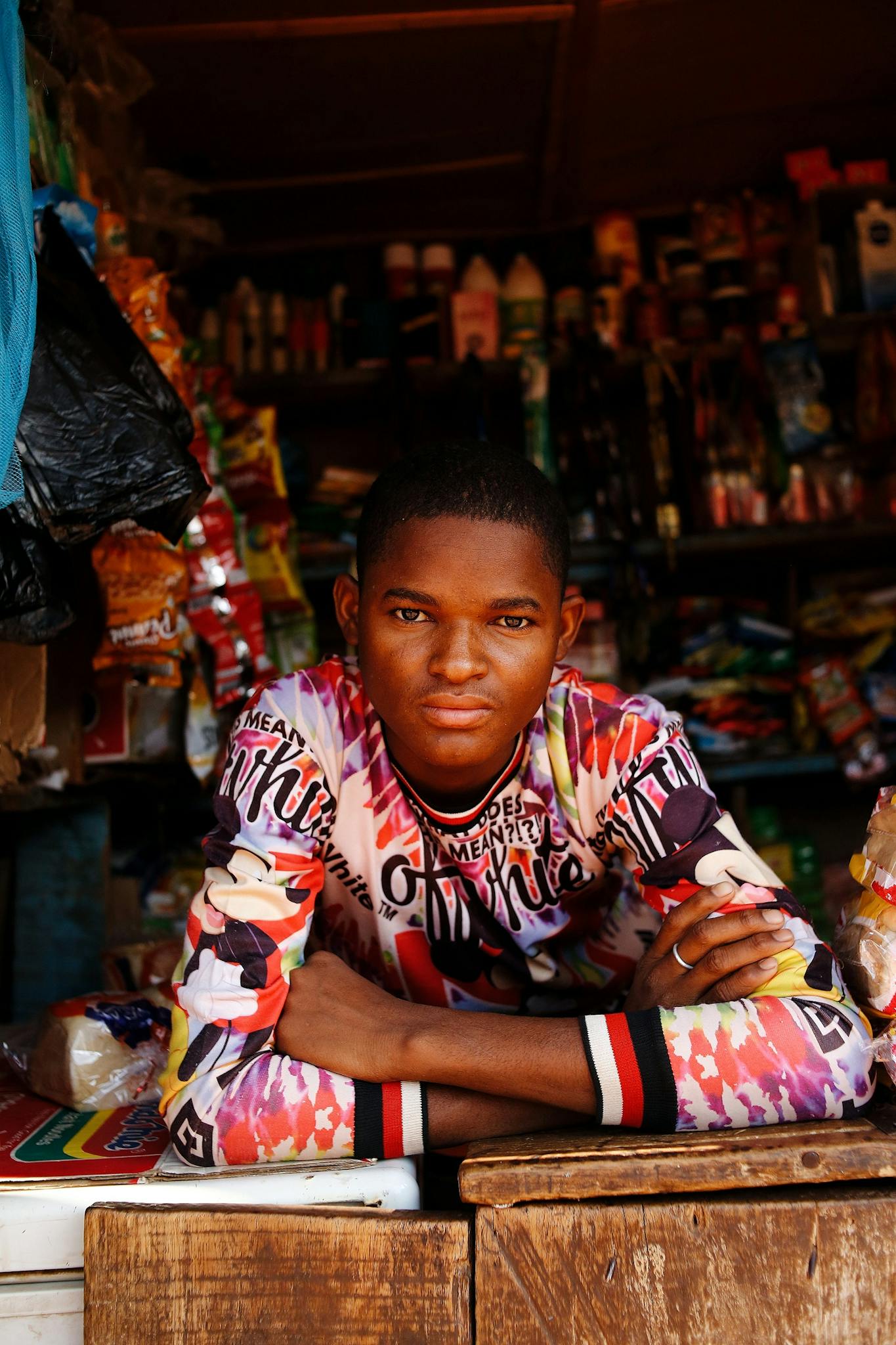 Portrait of a young man in a colorful shirt leaning on a Nigerian storefront counter.
